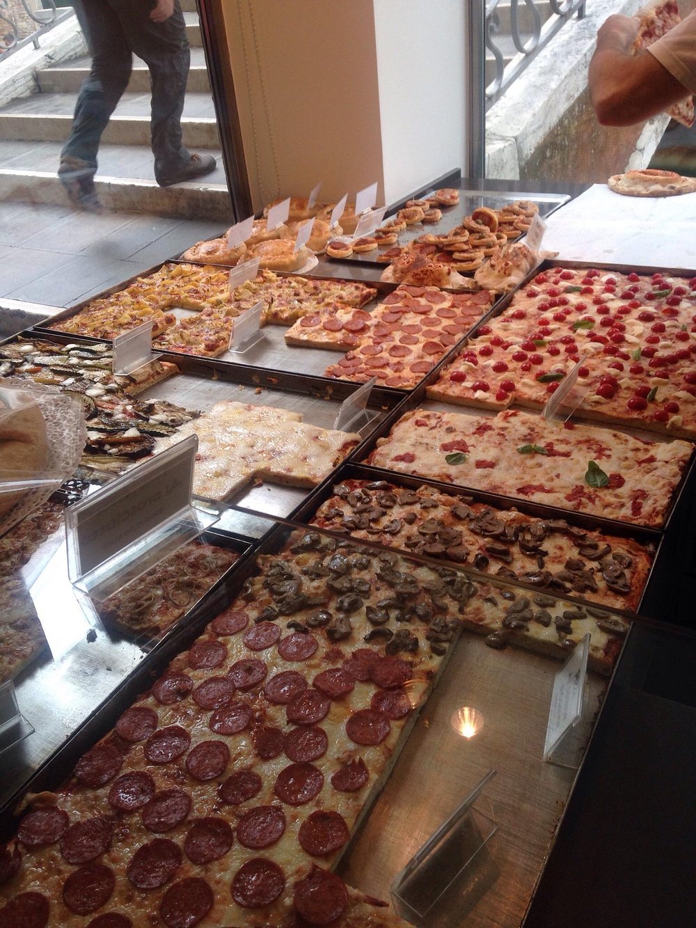 Teglie di pizza al trancio in vetrina con varietà di condimenti.